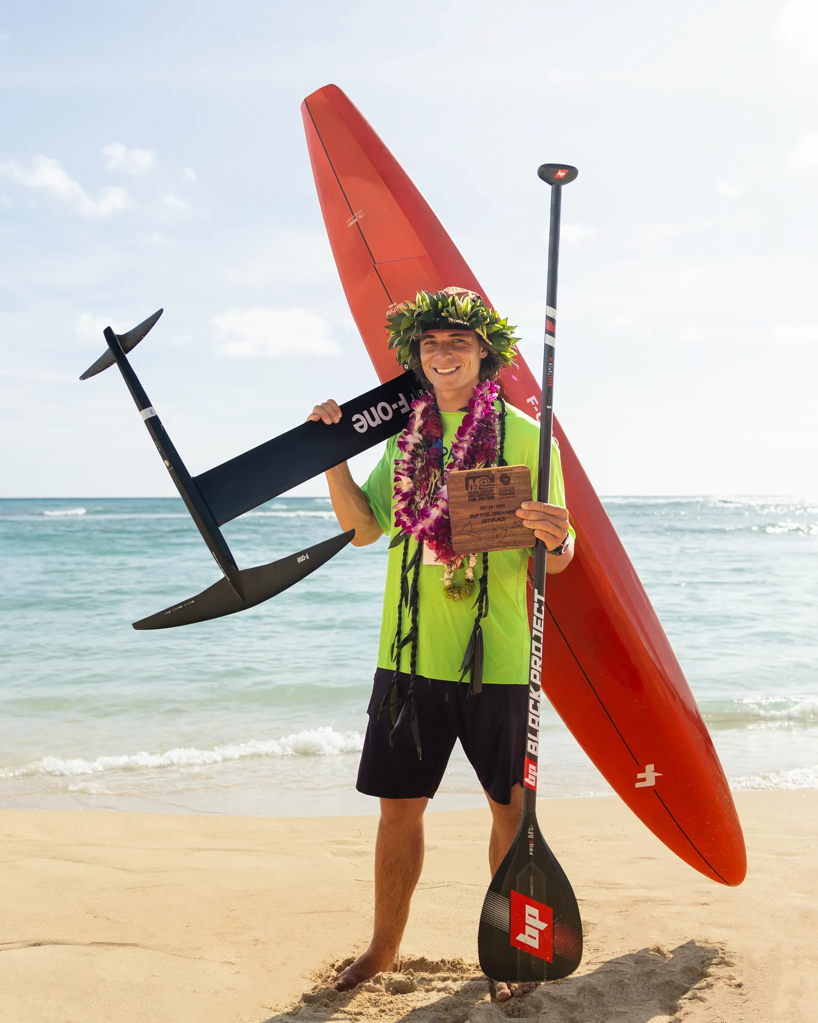 Edo Tanas on the beach with a lei, his foil board and paddle after a race win